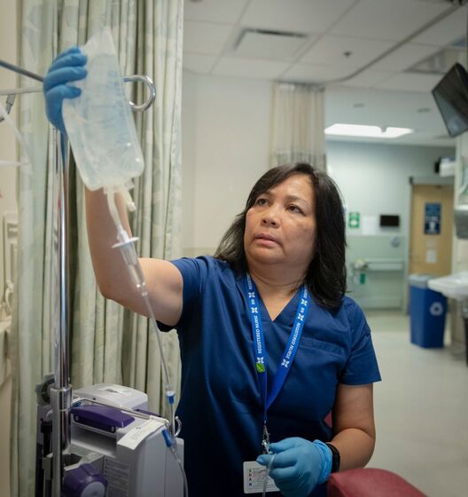 Maria Plummer in scrubs holding an IV bag in a hospital corridor